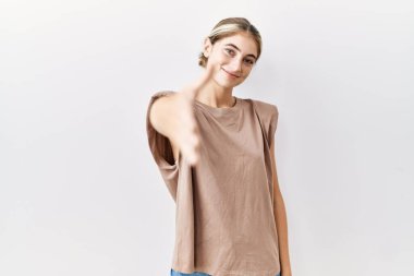 Young blonde woman standing over isolated background smiling friendly offering handshake as greeting and welcoming. successful business. 