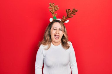 Orta yaşlı, beyaz bir kadın şirin Noel geyiği boynuzları takıyor kameraya seksi bir ifadeyle bakıyor, neşeli ve mutlu bir yüzle.. 