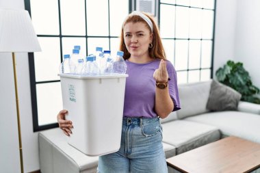 Elinde plastik şişelerle çöpleri geri dönüşüm sepetine koyan genç, kızıl saçlı bir kadın. Elleriyle para hareketi yapıyor. Maaş ödemesi istiyor. 