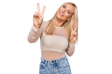 Sıradan kıyafetler giyen genç sarışın kadın kameraya bakıp zafer işareti yapıyor. İki numara.. 