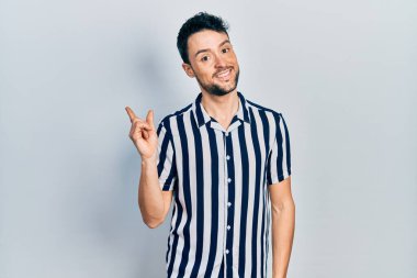 Yüzünde kocaman bir gülümseme olan, gündelik kıyafetler giyen genç İspanyol adam, kameraya el ve parmak ile bakıyor.. 