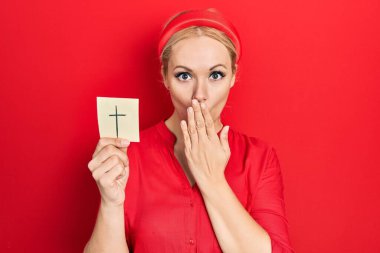 Young blonde woman holding catholic cross reminder covering mouth with hand, shocked and afraid for mistake. surprised expression 