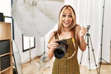 Young caucasian photographer girl holding professional camera at photography studio screaming proud, celebrating victory and success very excited with raised arms 
