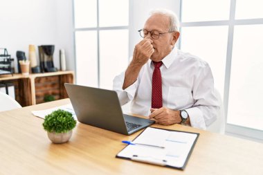 Senior man working at the office using computer laptop smelling something stinky and disgusting, intolerable smell, holding breath with fingers on nose. bad smell 