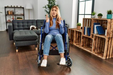 Young beautiful woman sitting on wheelchair at home crazy and mad shouting and yelling with aggressive expression and arms raised. frustration concept. 