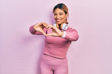 Güzel İspanyol kadın spor kıyafetleri giyiyor ve kulaklık takıyor. Sevgiyle gülümsüyor. Elleriyle kalp sembolü yapıyor. romantik kavram. 