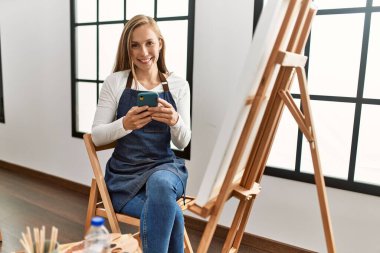 Beyaz kadın sanat stüdyosunda akıllı telefon kullanarak gülümsüyor.