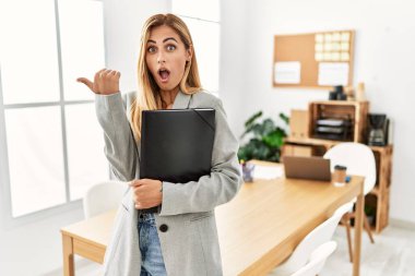 Blonde business woman at the office surprised pointing with hand finger to the side, open mouth amazed expression. 
