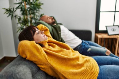 Genç İspanyol çift mutlu bir şekilde gülümsüyor. Elleri başının üstünde evdeki kanepede oturuyor..