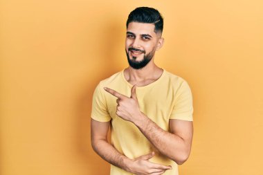 Sakallı, sıradan sarı tişörtlü yakışıklı, yüzünde mutlu ve doğal bir ifadeyle el ve parmak ile yüzünü gösteren bir gülümseme olan yakışıklı adam. 