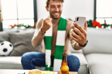 Genç İspanyol adam akıllı telefon kullanarak futbol maçını destekliyor evde Noel dekorunun yanında oturuyor.