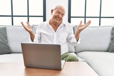 Senior man using laptop at home sitting on the sofa clueless and confused expression with arms and hands raised. doubt concept. 