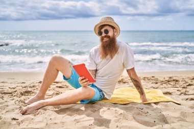 Genç kızıl saçlı adam plajda havlunun üzerinde oturuyor..