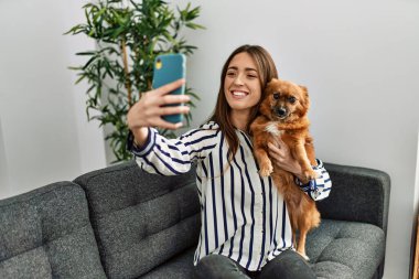 Genç İspanyol kadın akıllı telefonun başında selfie çekiyor. Kanepede oturup evdeki köpeğe sarılıyor.