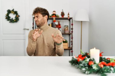 Young handsome man with beard sitting on the table by christmas decoration disgusted expression, displeased and fearful doing disgust face because aversion reaction. 