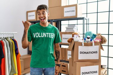 Young handsome hispanic man wearing volunteer t shirt at donations stand smiling with happy face looking and pointing to the side with thumb up. 