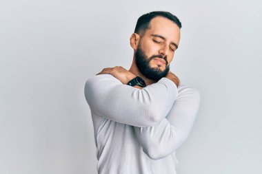 Sakallı genç adam, günlük beyaz gömlek giyiyor, kendine sarılıyor, mutlu ve pozitif, kendinden emin gülümsüyor. Sevgi ve özgüven. 