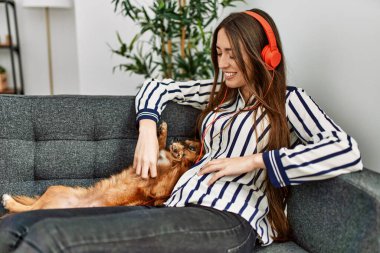 Young hispanic woman listening to music sitting on sofa with dog at home