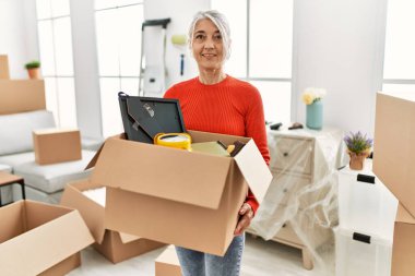Orta yaşlı, kır saçlı, gülümseyen bir kadın yeni evinde kendine güvenen bir paket tutuyor.