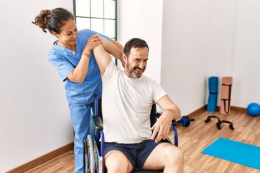 Orta yaşlı bir adam ve kadın, fizik tedavi kliniğinde tekerlekli sandalyeye bağlı olarak rehabilitasyon seansı yapıyorlar.