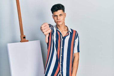 Genç İspanyol adam boş tuvale yakın duruyor mutsuz ve kızgın görünüyor reddediliyor ve olumsuz bir hareket yapıyor. kötü ifade. 