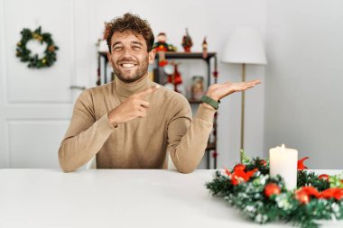Young handsome man with beard sitting on the table by christmas decoration amazed and smiling to the camera while presenting with hand and pointing with finger. 