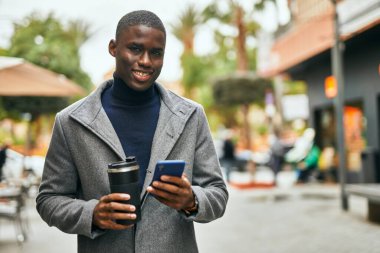 Genç Afrikalı Amerikalı adam akıllı telefon kullanıyor. Şehirde kahve içiyor.