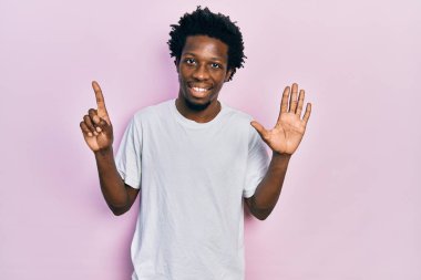 Genç Afro-Amerikan adam, günlük beyaz tişört giyip altı numaralı parmaklarıyla işaret ederken kendinden emin ve mutlu gülümsüyor.. 