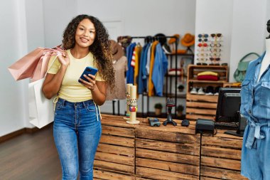 Genç Latin müşteri kadın gülümseyerek alışveriş torbaları tutuyor ve giyim mağazasında akıllı telefon kullanıyor..