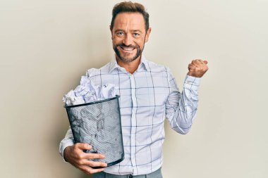 Orta yaşlı bir adam elinde buruşuk kağıtlarla dolu bir kağıt kutuyla başparmağıyla yan tarafa bakıp mutlu bir şekilde gülümsüyor. 