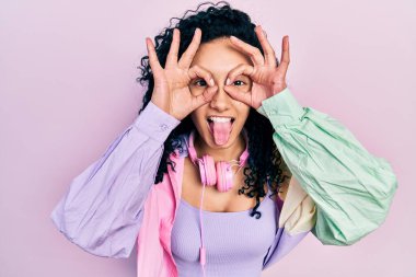 Young hispanic woman with curly hair wearing gym clothes and using headphones doing ok gesture like binoculars sticking tongue out, eyes looking through fingers. crazy expression. 