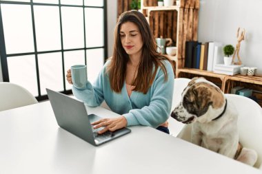 Evde köpekle aynı masada oturan dizüstü bilgisayar kullanan genç bir kadın.