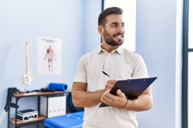 Fizyoterapist üniforması giyen genç İspanyol adam klinikteki panoya yazı yazıyor.