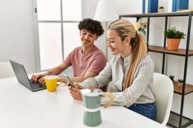 Laptop ve akıllı telefon kullanan genç bir çift evde kahve içiyor..
