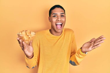 Genç Afrikalı Amerikalı adam patates cipsi tutuyor başarıyı mutlu bir gülümsemeyle kutluyor ve kazanan el kaldırarak ifade ediyor 