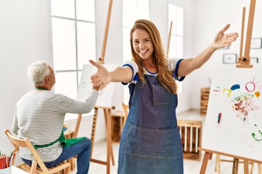 Hispanic woman wearing apron at art studio looking at the camera smiling with open arms for hug. cheerful expression embracing happiness. 