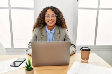 Ofisteki masasında oturan güzel İspanyol iş kadını yüzünde mutlu ve havalı bir gülümsemeyle dizüstü bilgisayarla çalışıyor. şanslı kişi. 