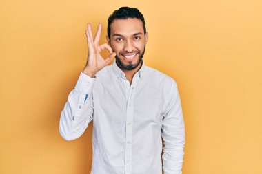 Sakallı, iş gömleği giyen, gülümseyen, el ve parmaklarıyla imza atan İspanyol bir adam. başarılı ifade. 