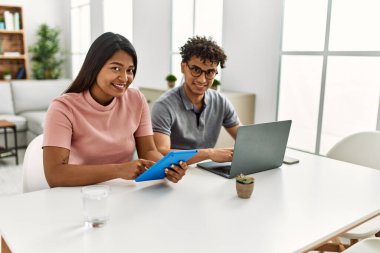 Genç latin çift evde masanın üzerinde dizüstü bilgisayar ve dokunmatik ped kullanarak çalışıyor..