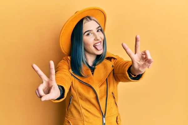 黄色の帽子と革のジャケットを身に着けている若い現代の女の子は 両方の手の指が勝利のサインを行うことを示す舌で笑顔 — ストック写真
