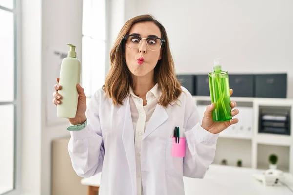 Young doctor woman holding aloe vera and cream making fish face with mouth and squinting eyes, crazy and comical.