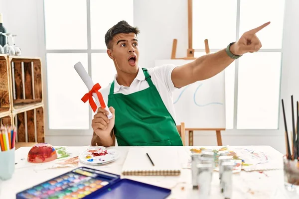 Young Hispanic Man Art Studio Holding Degree Pointing Finger Surprised — стоковое фото