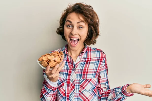 Mujer Morena Joven Sosteniendo Tazón Con Galletas Saladas Celebrando Logro —  Fotos de Stock