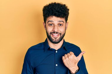 Young arab man with beard wearing casual shirt smiling with happy face looking and pointing to the side with thumb up. 
