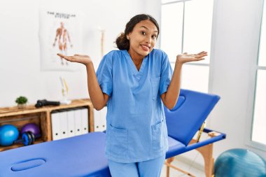Güzel İspanyol fizyoterapist kadın ağrı kesici kliniğinden habersiz ve elleri ve kolları havada kafası karışmış bir ifade. şüphe kavramı. 