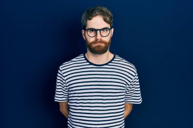 Sakallı, çizgili tişörtlü, gözlüklü, kuşkucu ve sinirli bir beyaz adam sorun yüzünden surat asıyor. negatif kişi. 