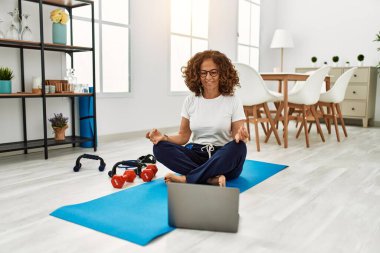Orta yaşlı İspanyol kadın evde dizüstü bilgisayar kullanarak yoga eğitimi alıyor.