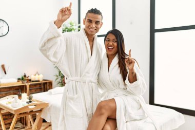 Young latin couple wearing towel standing at beauty center pointing finger up with successful idea. exited and happy. number one. 