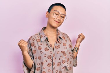 Kısa saçlı, güzel İspanyol bir kadın. Gözlüklü, mutlu ve heyecanlı. Kolları havada, gülümseyen ve başarı için bağıran, kazanan bir jest yapıyor. kutlama konsepti. 