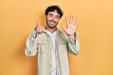 Latin asıllı genç adam gündelik giysiler giyip altı numaralı parmaklarıyla işaret ederken kendinden emin ve mutlu bir şekilde gülümsüyor.. 